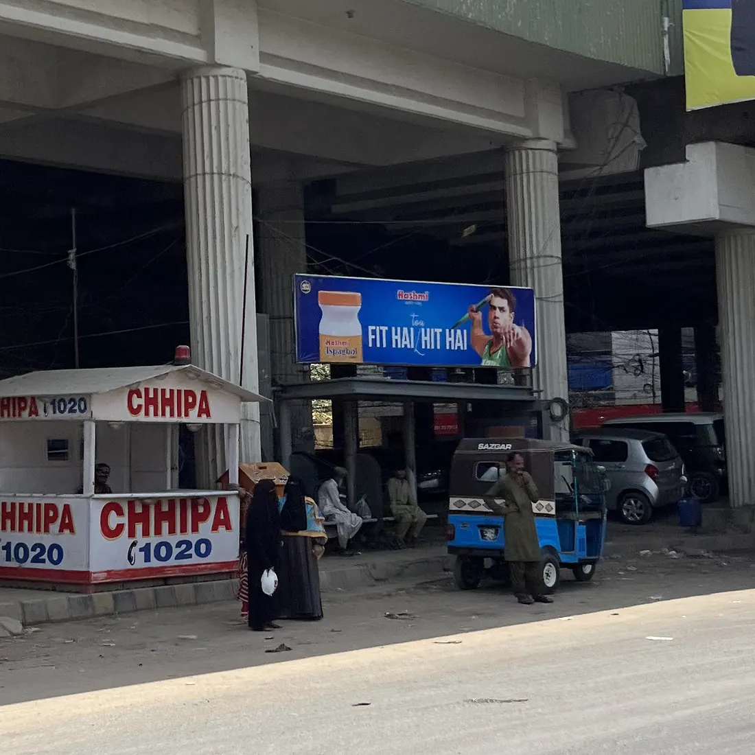 Waiting beneath a flyover: men sit on a bench in the shade while two women stand apart in the open, beside a first-aid booth and a rickshaw.