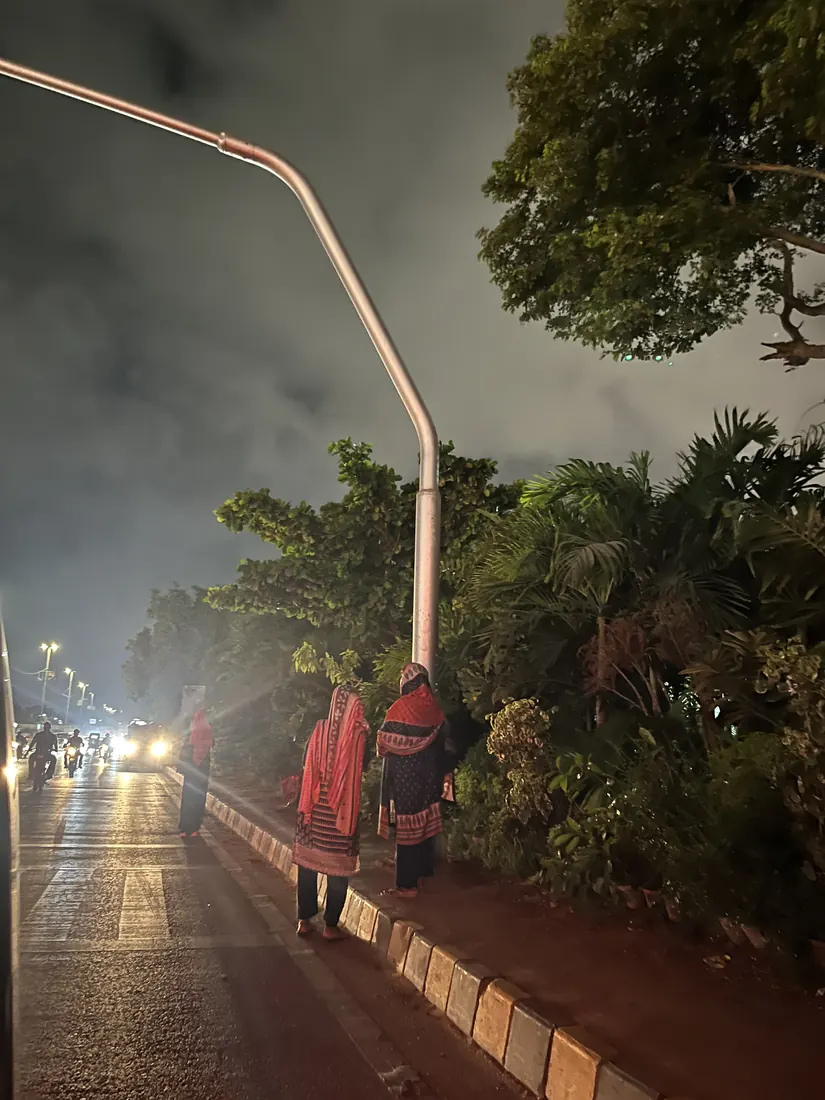 Two women wait at an unlit roadside kerb at night, lit only by the headlights of approaching traffic.