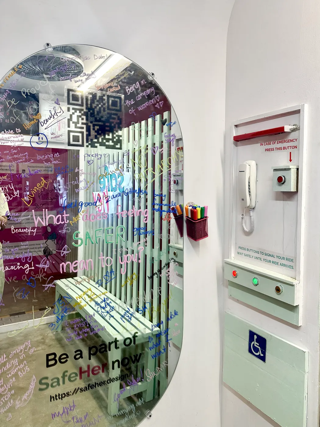 Inside the SafeStop: the prompt 'What does SafeHer mean to you?' answered in coloured marker by visitors, beside the helpline panel, accessible seating and a QR code inviting people to join.